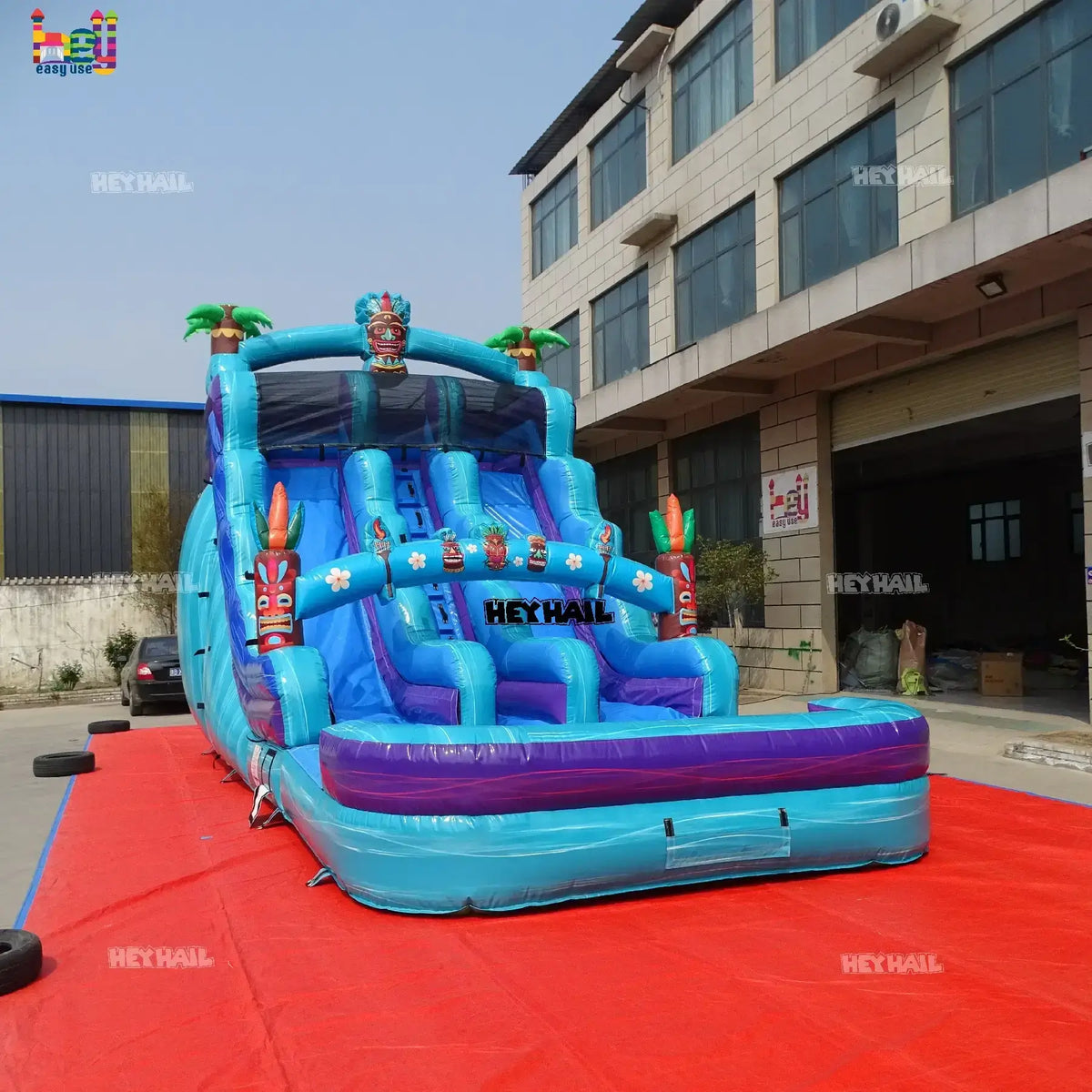 Outdoor three-quarter view of a teal and blue 18ft dual-lane inflatable water slide with tiki mask, palm-tree accents, and a landing pool
