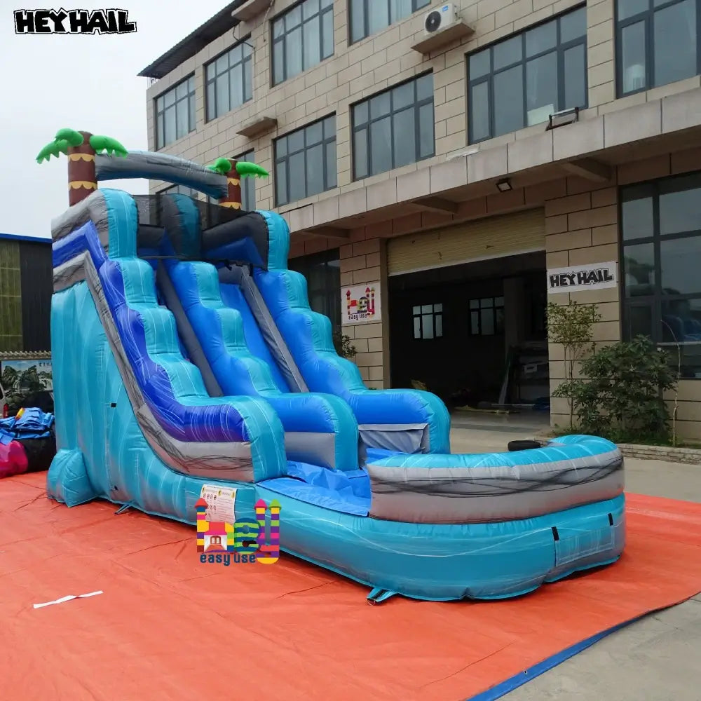 Outdoor angled front view of a marble-blue single-lane inflatable water slide with a gray landing pool and palm-tree arch