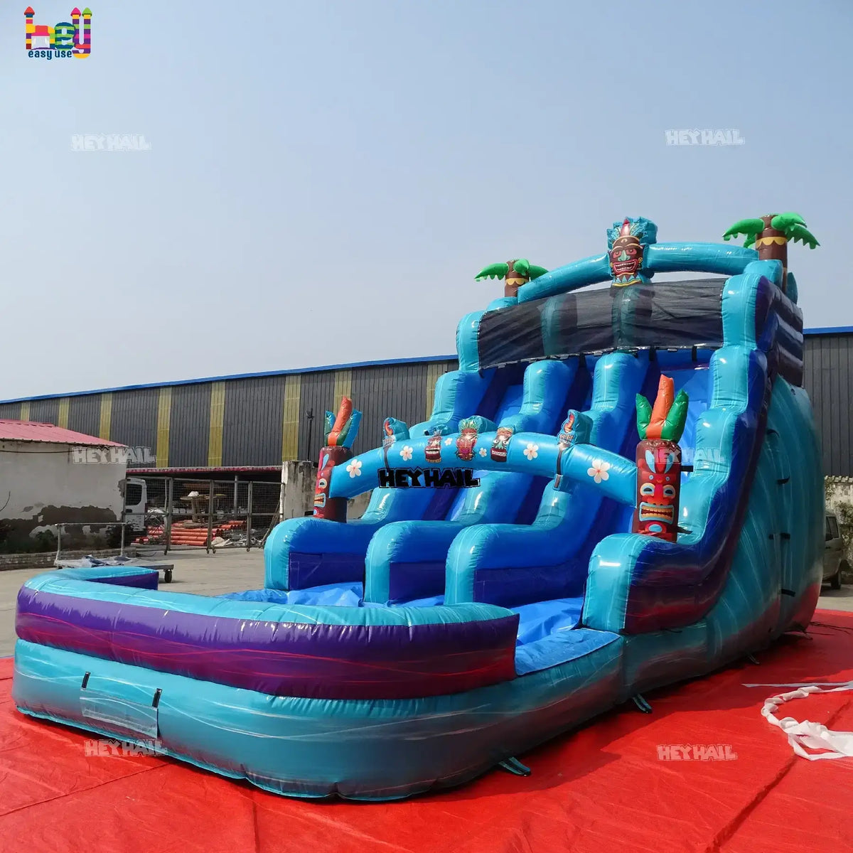Outdoor angled view of a teal and blue 18ft dual-lane inflatable water slide with tiki mask, palm-tree accents, and a landing pool