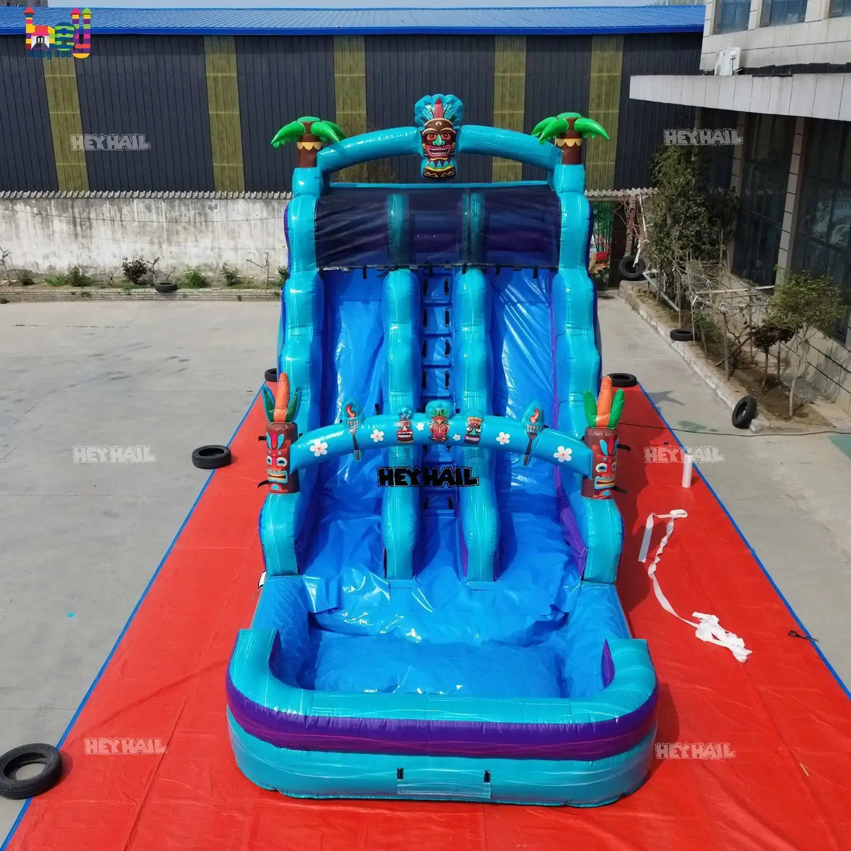 Outdoor front view of a teal and blue 18ft dual-lane inflatable water slide with tiki mask, palm-tree accents, and a landing pool