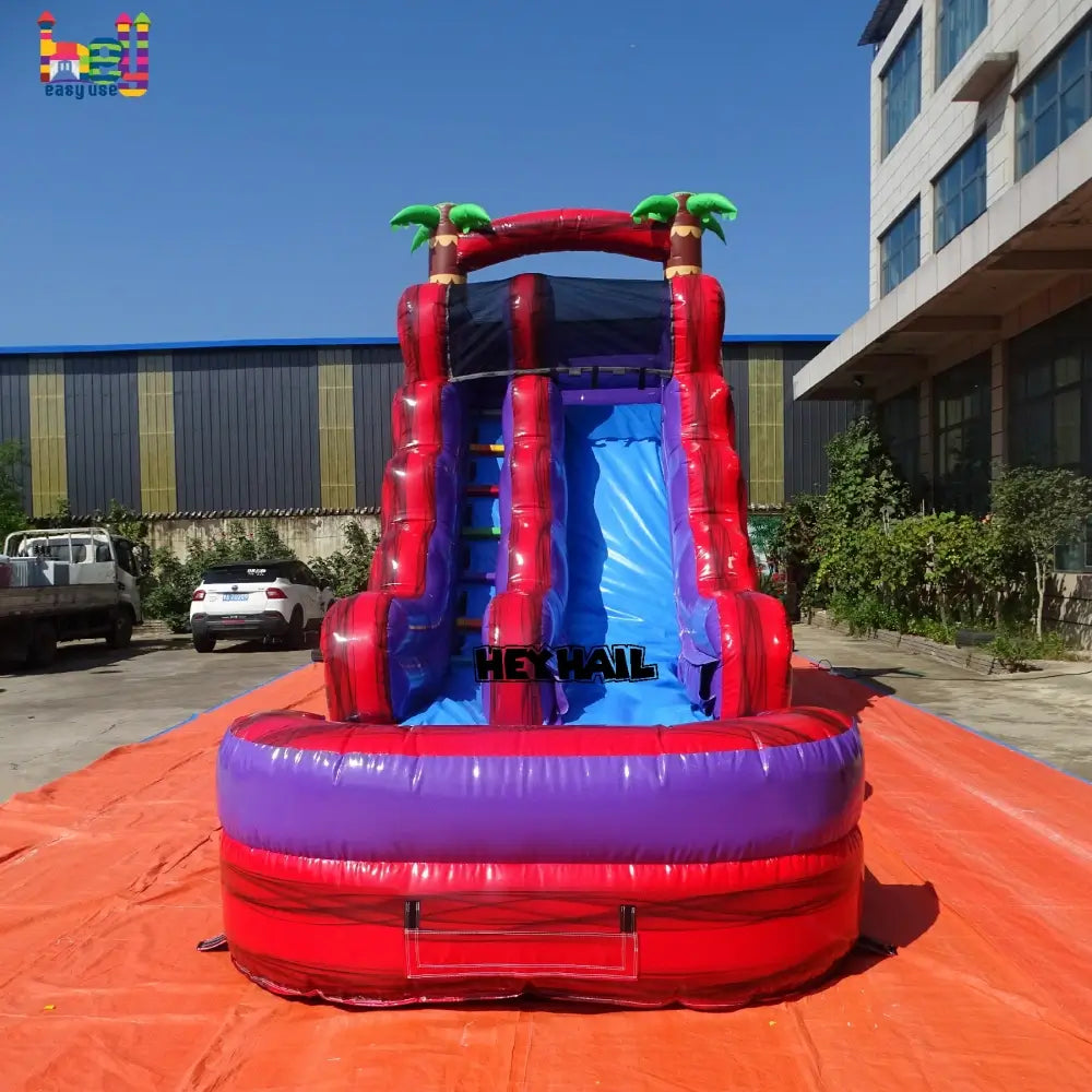 Outdoor front view of red-and-purple front-load water slide with blue slide lane, attached landing pool, and palm-tree accents